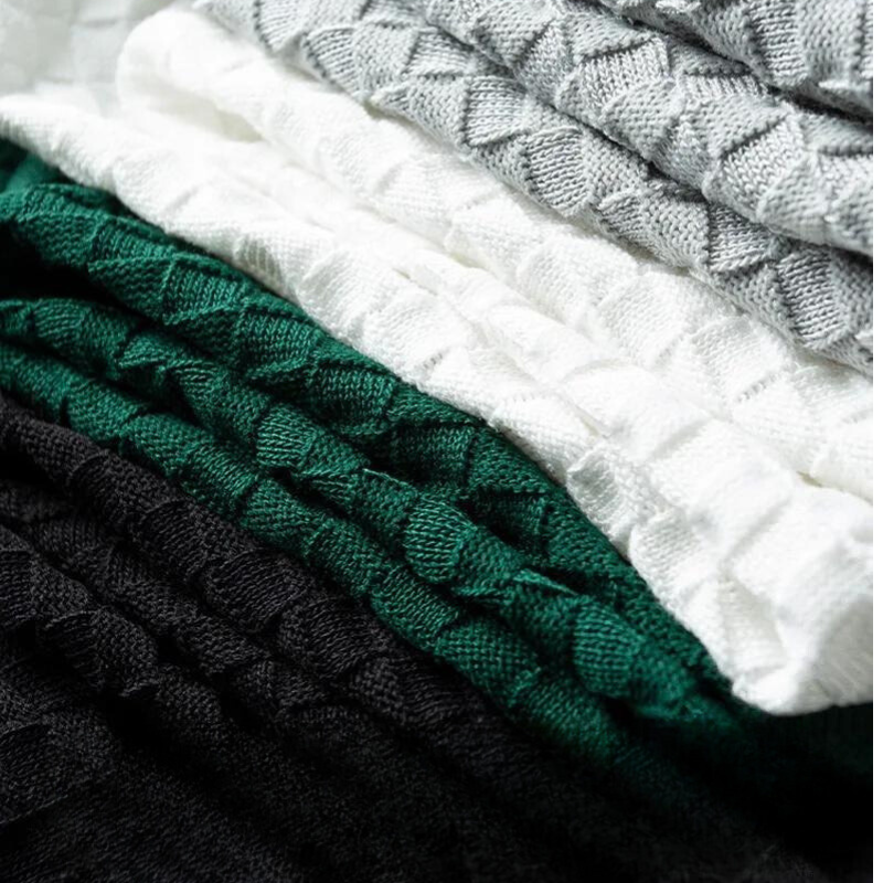 A close-up of neatly folded knit sweaters in black, dark green, white, and light gray, featuring textured patterns like waffle jacquard and showcasing soft, cozy fabric.