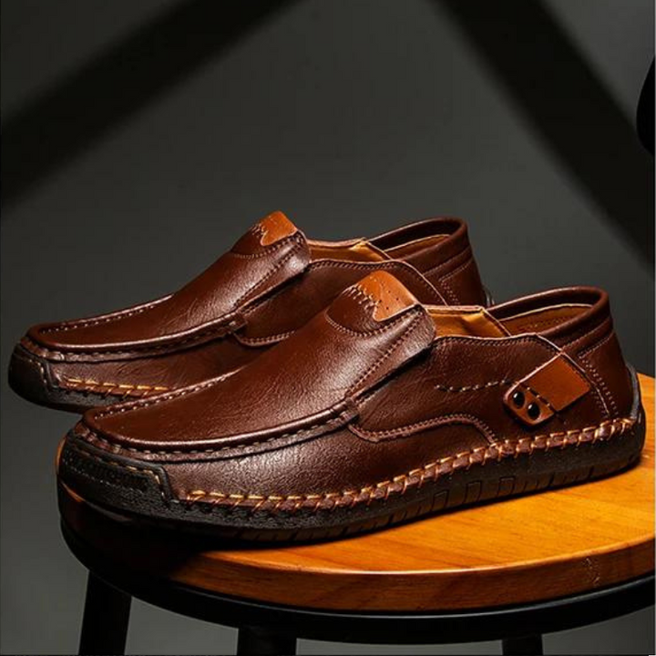 A pair of brown leather loafers with detailed stitching and textured rubber soles, placed side by side on a wooden stool against a dark background.