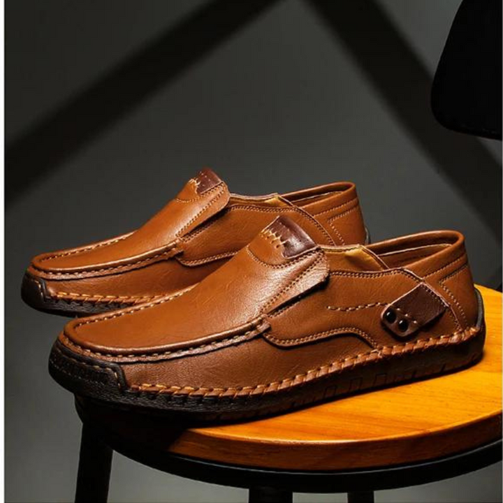 A pair of brown leather loafers with detailed stitching and textured soles, displayed on a wooden stool under dramatic lighting, showcases traditional craftsmanship.