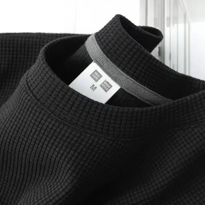 A close-up of a black, waffle-textured shirt with a gray inner collar and a white size tag labeled GU Dry and M for medium. The breathable shirts fabric detail and neckline are clearly visible.