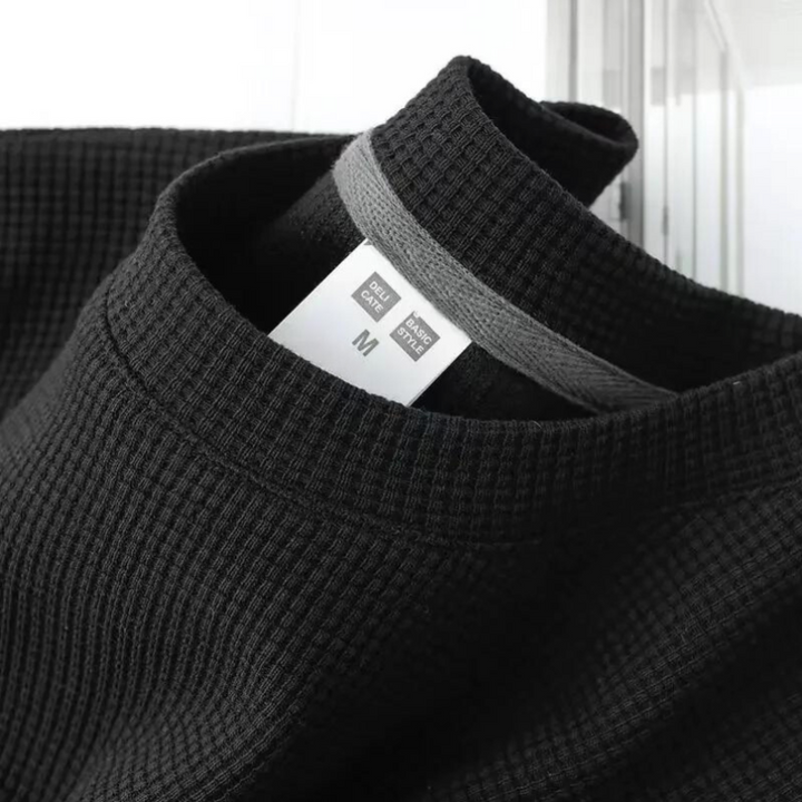 A close-up of a black, waffle-textured shirt with a gray inner collar and a white size tag labeled GU Dry and M for medium. The breathable shirts fabric detail and neckline are clearly visible.