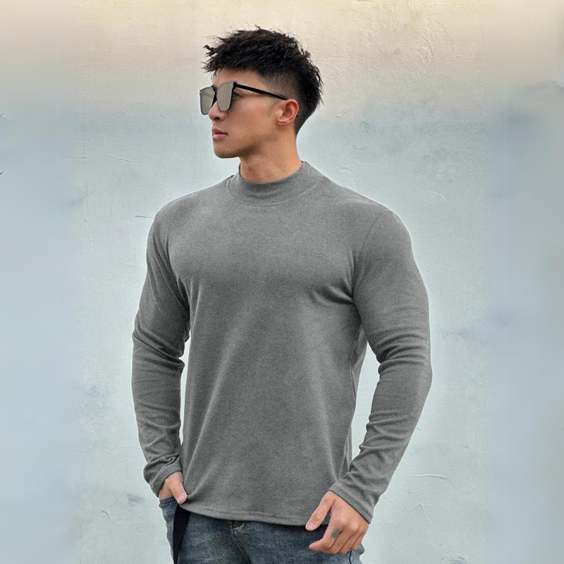 A man wearing large sunglasses, a fitted gray long-sleeve shirt with a mock neck collar, and jeans stands against a light gray textured background, gazing to his left with one hand in his pocket.