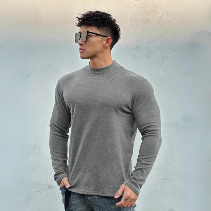 A man wearing large sunglasses, a fitted gray long-sleeve shirt with a mock neck collar, and jeans stands against a light gray textured background, gazing to his left with one hand in his pocket.