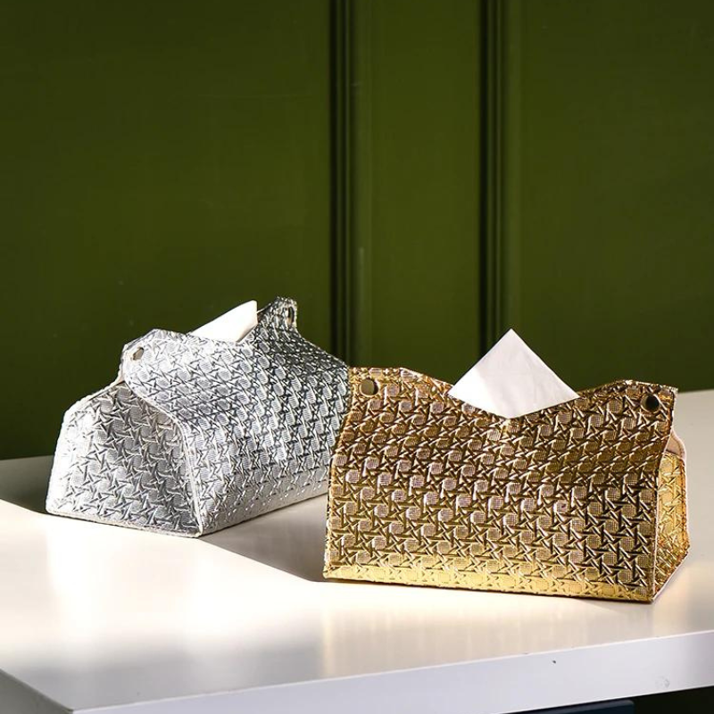 Two decorative tissue holders, the Glymmer Tissue Box in metallic silver and gold, sit on a white surface against a dark green background. Both feature a striking geometric pattern and hold tissues.