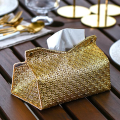 A gold, textured Glymmer Tissue Box with a geometric pattern sits on a wooden table, surrounded by gold cutlery, a glass, and plates, creating an elegant dining setting.
