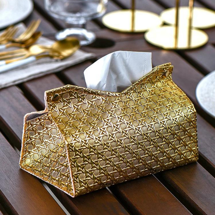 A gold, textured Glymmer Tissue Box with a geometric pattern sits on a wooden table, surrounded by gold cutlery, a glass, and plates, creating an elegant dining setting.