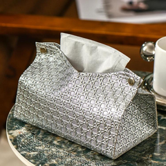 A silver, textured Glymmer Tissue Box with a geometric pattern sits on a marble table next to a white cup and saucer. A sheet of tissue is partially pulled from this decorative tissue holder.