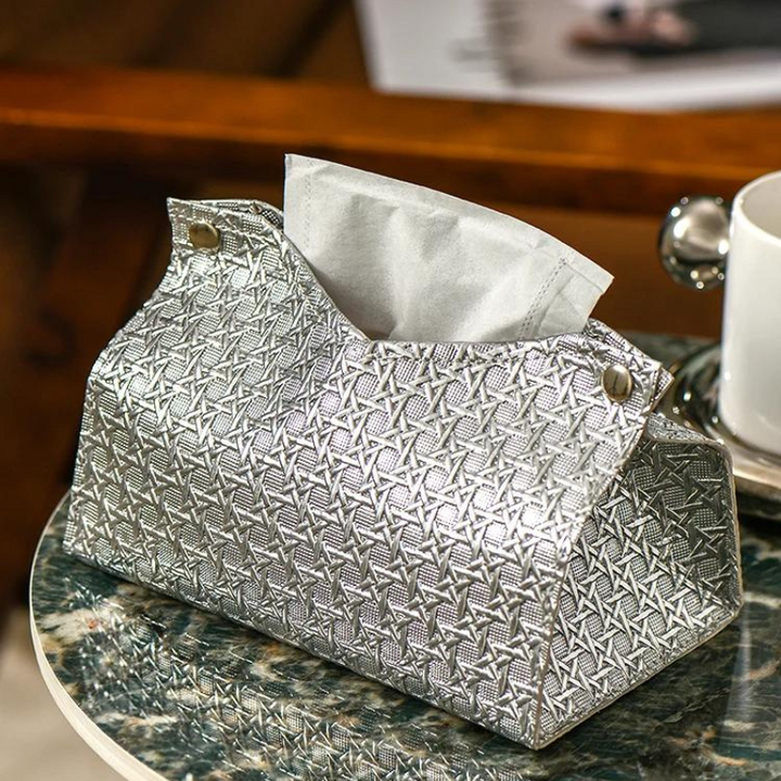 A silver, textured Glymmer Tissue Box with a geometric pattern sits on a marble table next to a white cup and saucer. A sheet of tissue is partially pulled from this decorative tissue holder.