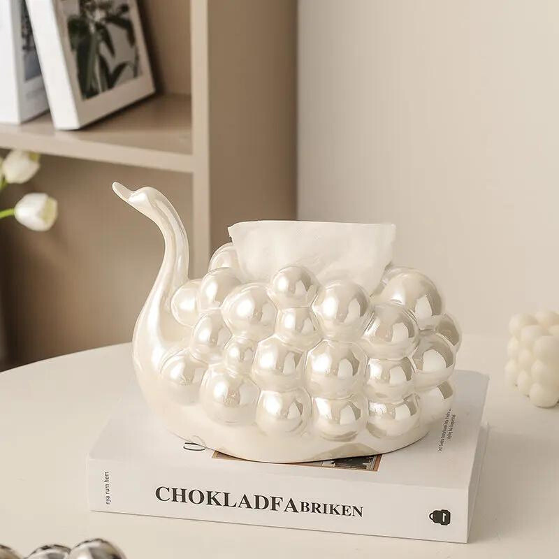 A pearl white dispenser shaped like a swan with a rounded, bulbous body sits on a book titled CHOKLADFABRIKEN on a light-colored table. A tissue is partially pulled from the elegant swan design.