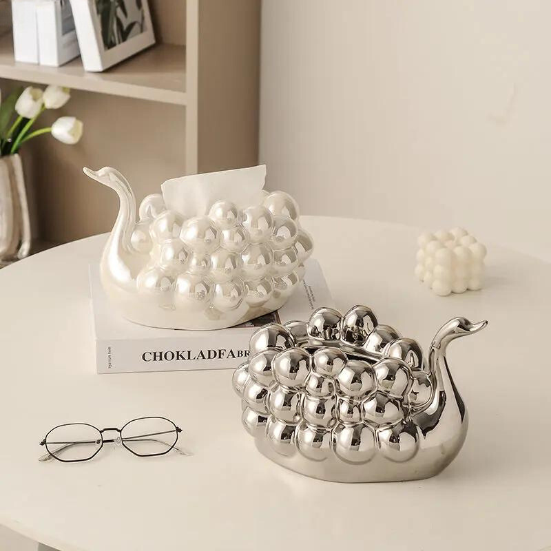 Two decorative snail-shaped containers with a bumpy, shiny surface sit on a table; one is a pearl white dispenser for tissues. Nearby are a pair of glasses, a small cube candle, a flower vase, and a book on the shelf in the background.