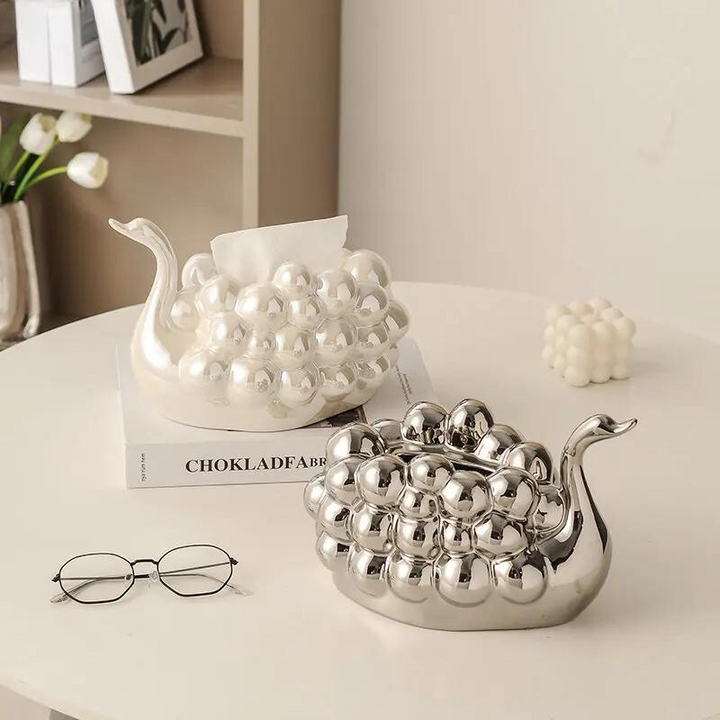 Two decorative snail-shaped containers with a bumpy, shiny surface sit on a table; one is a pearl white dispenser for tissues. Nearby are a pair of glasses, a small cube candle, a flower vase, and a book on the shelf in the background.