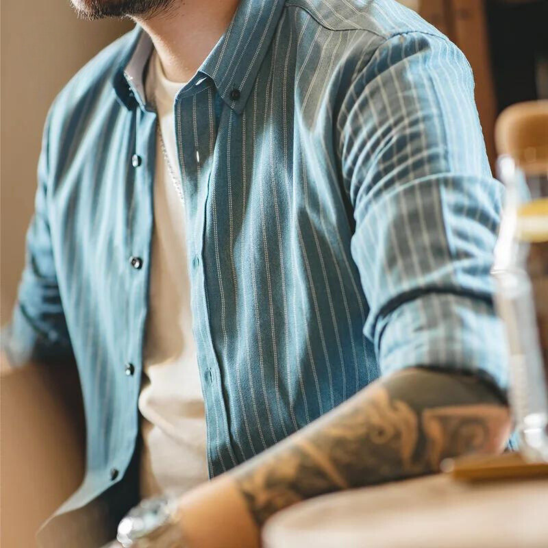 A person wearing a blue pinstripe shirt made from fine weave cotton over a white t-shirt sits at a table. Their arms, showing a watch and tattoos, rest on the tabletop. The image is cropped to show only the torso and arms.