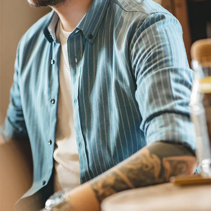 A person wearing a blue pinstripe shirt made from fine weave cotton over a white t-shirt sits at a table. Their arms, showing a watch and tattoos, rest on the tabletop. The image is cropped to show only the torso and arms.