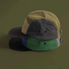 Two five-panel patchwork caps are stacked against an olive green background, channeling an earthtone aesthetic; the top cap blends tan, gray, and black, while the bottom features green, gray, blue, and black sections.