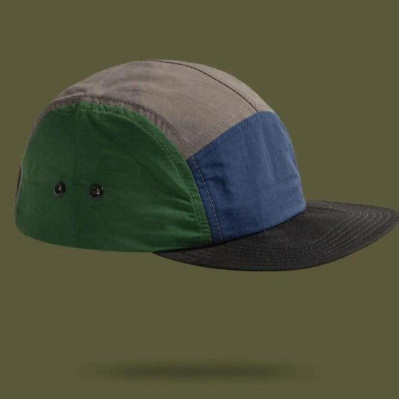 A color-blocked patchwork cap with green, gray, and blue panels, featuring a black brim and two small ventilation holes on the green side panel, set against an olive green background for an earthtone aesthetic.