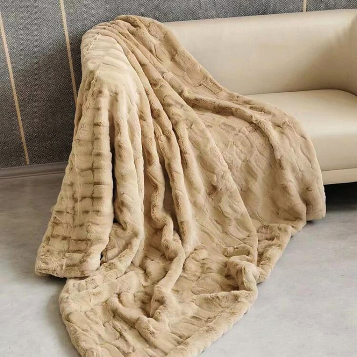 A large, plush faux fur blanket in soft beige is draped over a light-colored sofa, cascading to the floor in gentle folds. The background features a grey wall with subtle vertical accents.