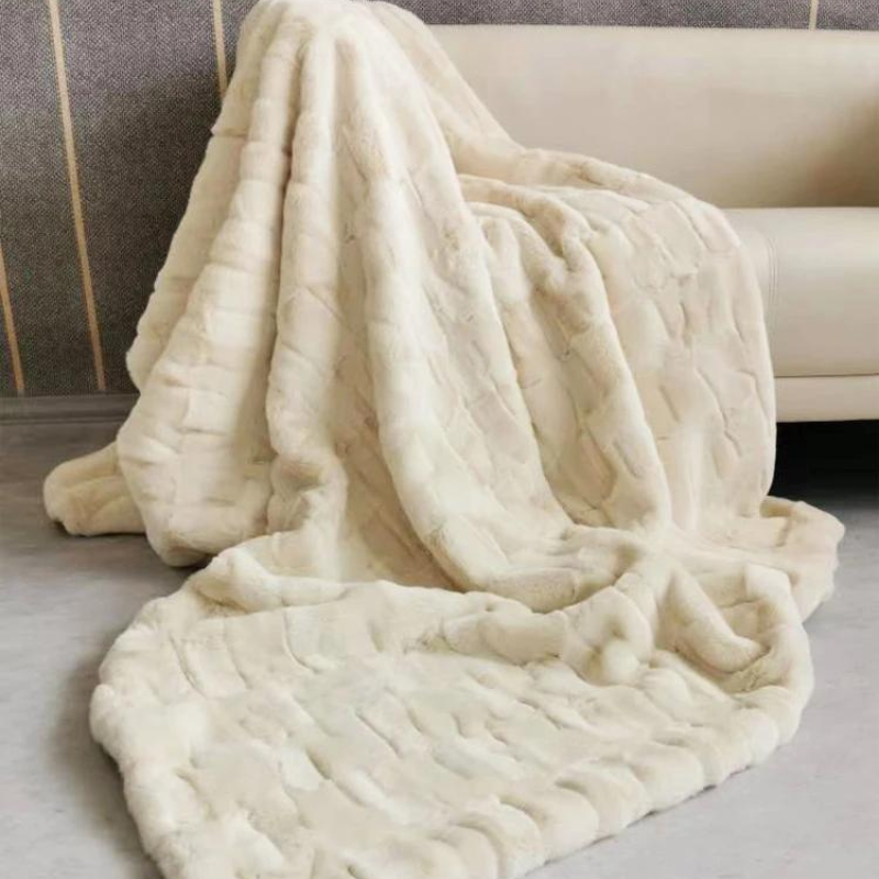 A plush faux fur, cream-colored blanket drapes over the side of a light-colored sofa, spilling onto the floor in soft folds. The eco-friendly blanket features a textured, quilted pattern for added coziness.