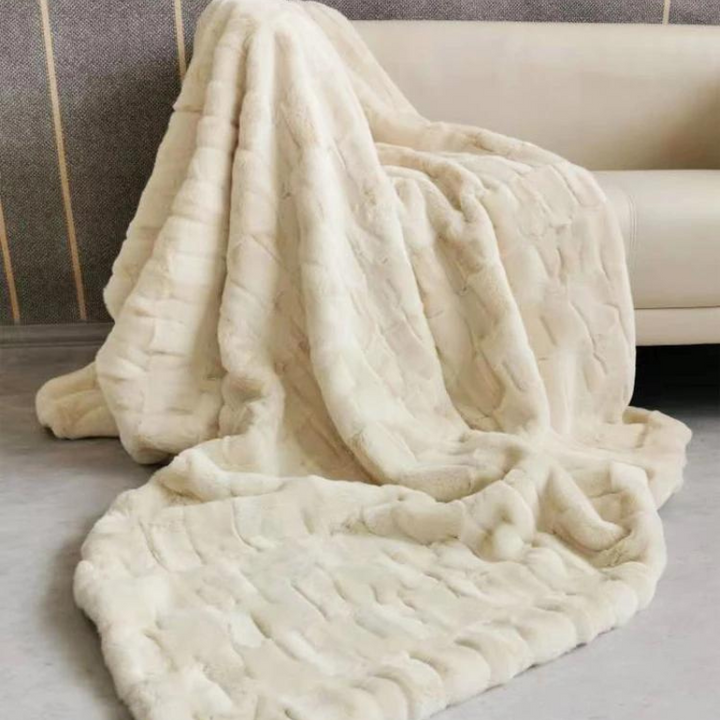 A plush faux fur, cream-colored blanket drapes over the side of a light-colored sofa, spilling onto the floor in soft folds. The eco-friendly blanket features a textured, quilted pattern for added coziness.