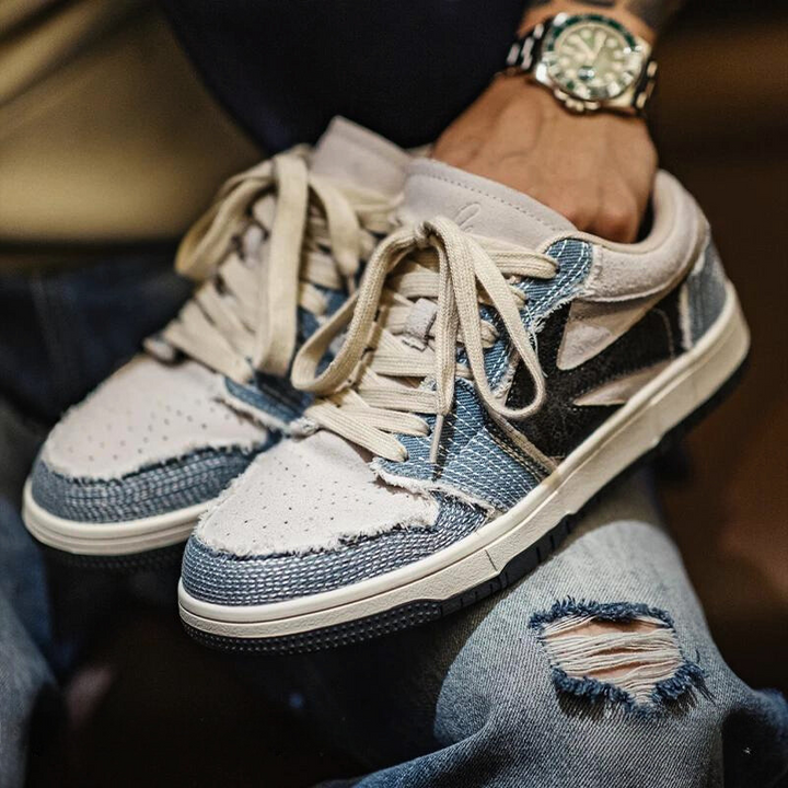 A person wearing a watch holds a pair of Denimwave 1986 Pro Sneakers with frayed edges and EVA midsole cushioning, while wearing ripped blue jeans for an effortlessly cool 80s aesthetic.