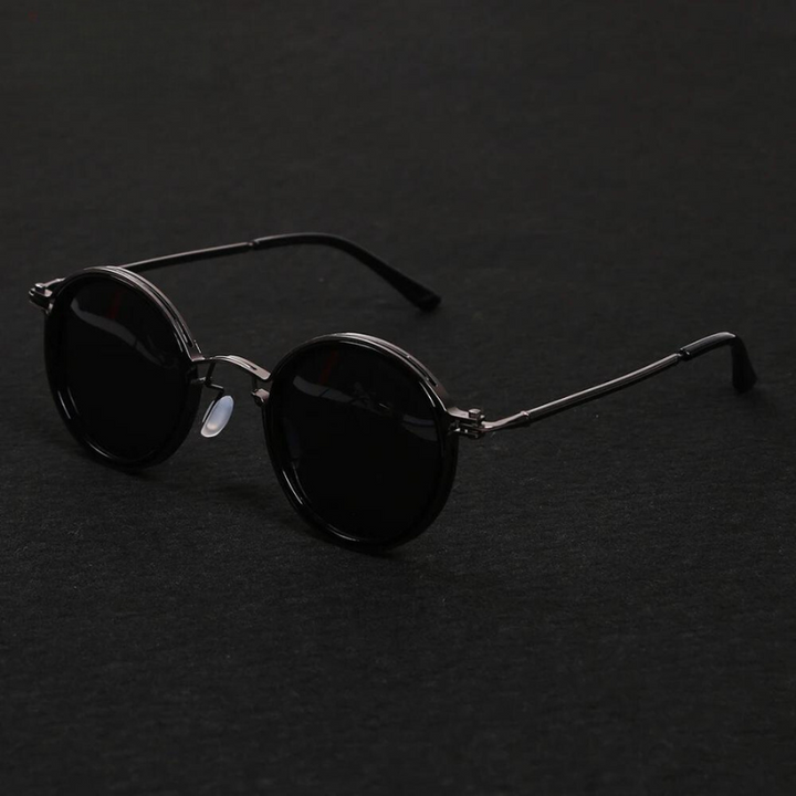 Round black Ruby Ray Sunglasses with dark lenses and thin metal frames are placed on a dark surface, their arms extended. These polarized sunglasses offer both style and enhanced UV400 protection for your eyes.