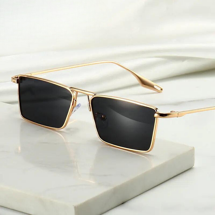A pair of stylish Avalon Sunglasses featuring gold frames and rectangular lenses rests on a white marble surface against a soft, light-colored background.