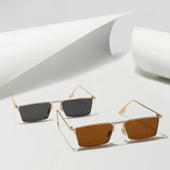 Two pairs of Avalon Sunglasses with gold frames and rectangular lenses rest on a white surface—one with dark gray lenses, the other with brown. Rolled pieces of white paper are in the background.