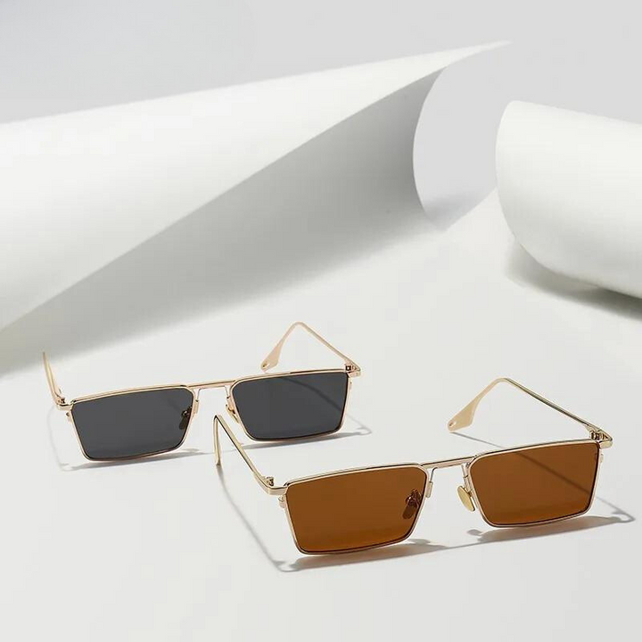 Two pairs of Avalon Sunglasses with gold frames and rectangular lenses rest on a white surface—one with dark gray lenses, the other with brown. Rolled pieces of white paper are in the background.