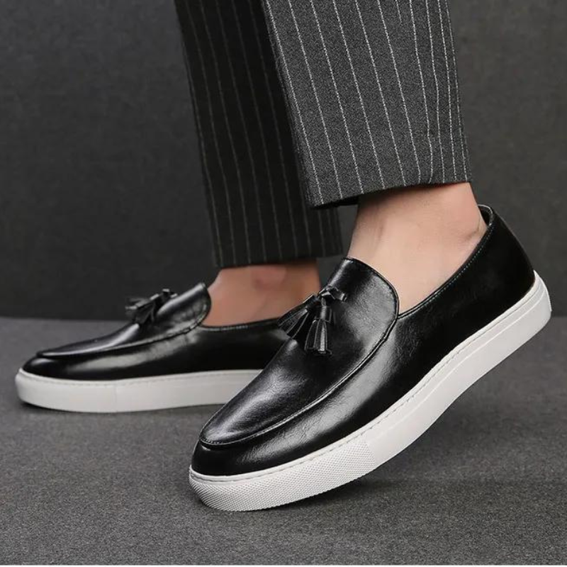 A person wearing luxury leather tassel loafers with white rubber soles and cushioned insoles, paired with dark pinstripe trousers, stands on a grey surface.