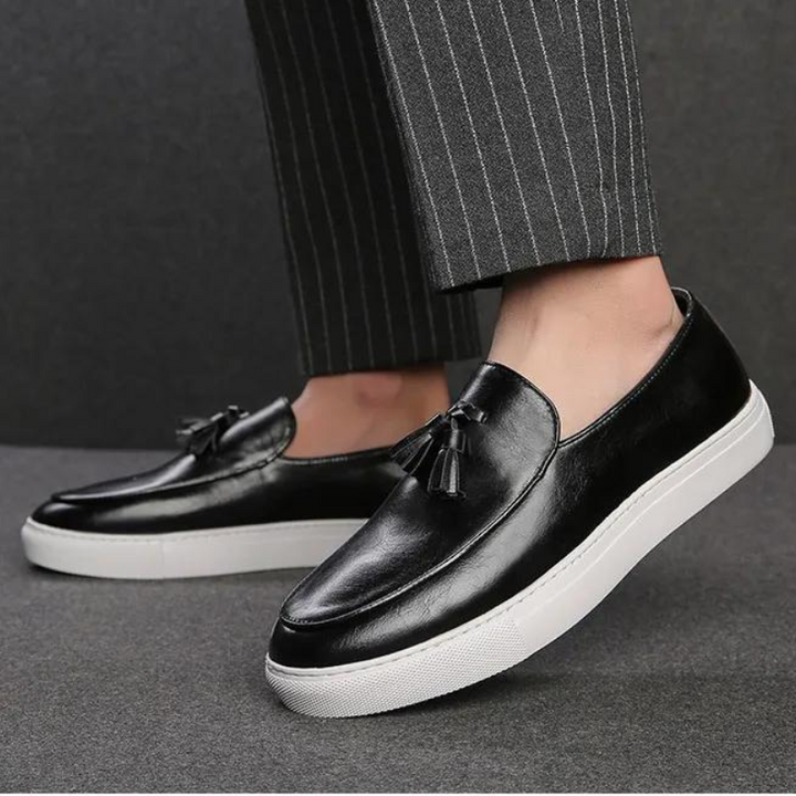 A person wearing luxury leather tassel loafers with white rubber soles and cushioned insoles, paired with dark pinstripe trousers, stands on a grey surface.