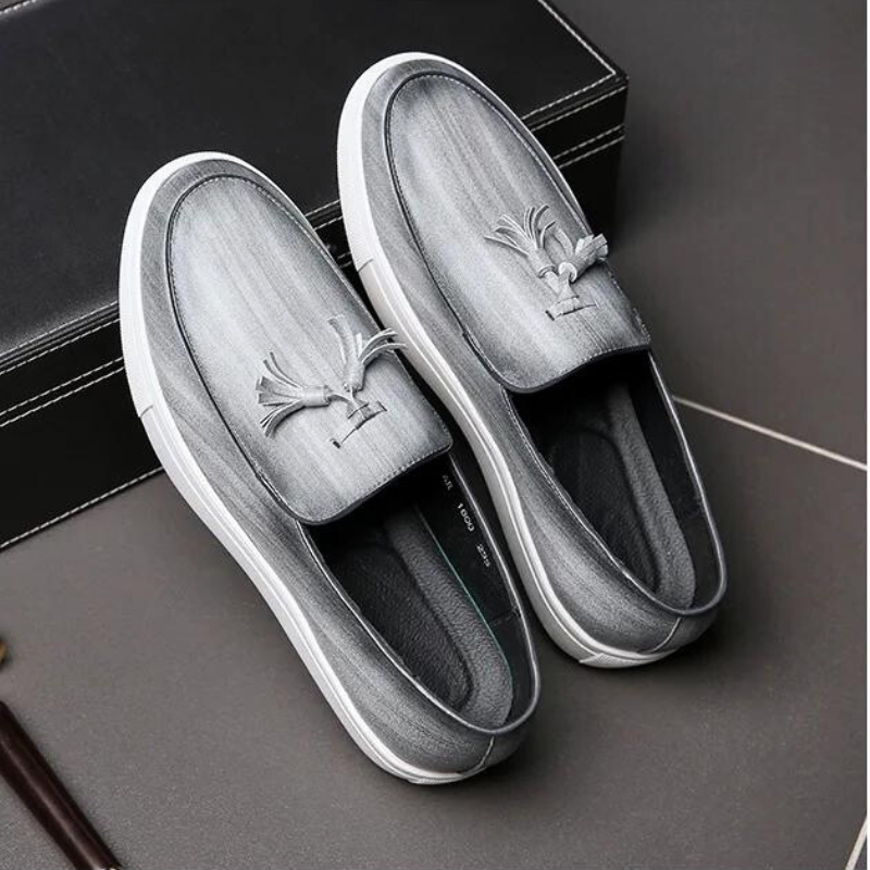 A pair of stylish gray tassel loafers with white soles and decorative tassels on top, featuring a cushioned insole for comfort, displayed next to a black box on a dark surface.