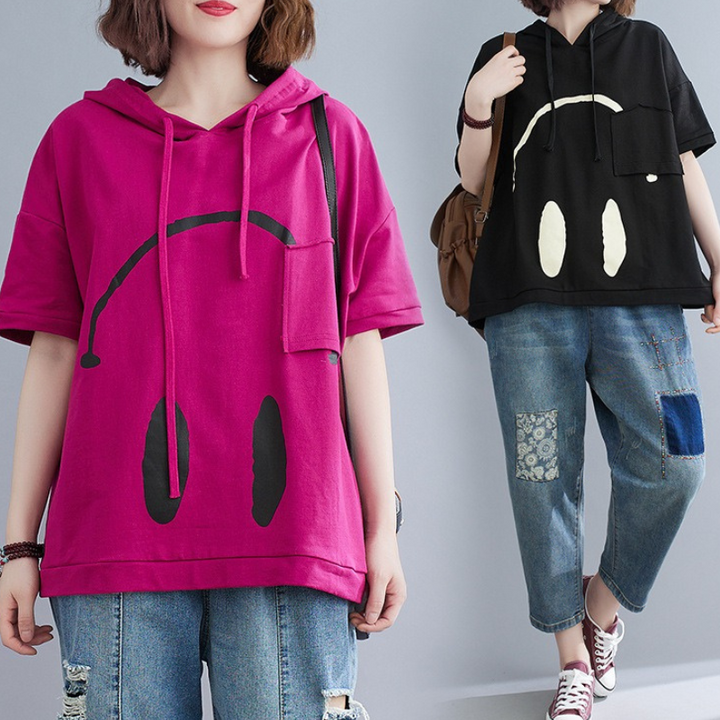 Two women wear loose-fit Monday Mood hoodies with a playful face design—one in magenta and one in black. Both pair these short-sleeve hoodies with baggy jeans and casual shoes against a plain background.