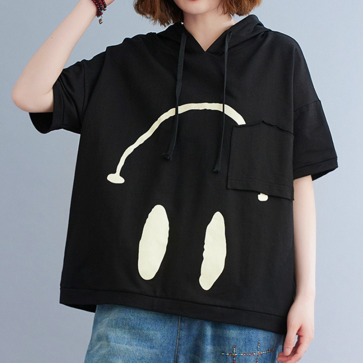 A person wearing a loose black short-sleeve hoodie with a simple white smiley face design, paired with blue denim jeans. The face is partially obscured by the pocket on this playful Monday Mood hoodie.