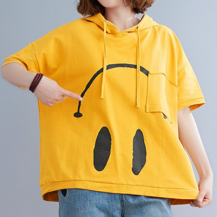 A person wearing a loose yellow Monday Mood hoodie with a large black abstract smiley face design points to the print. Only their torso is visible; they are also wearing blue jeans.