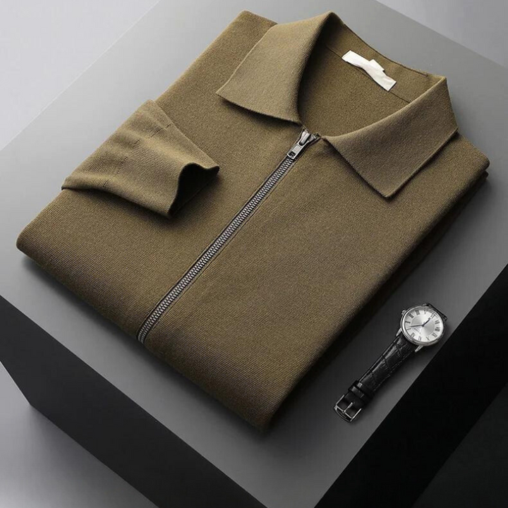 A folded olive green merino wool zip-up collared shirt with one sleeve partially visible sits on a gray surface next to a black leather strap wristwatch with a silver face, offering natural temperature regulation and style.