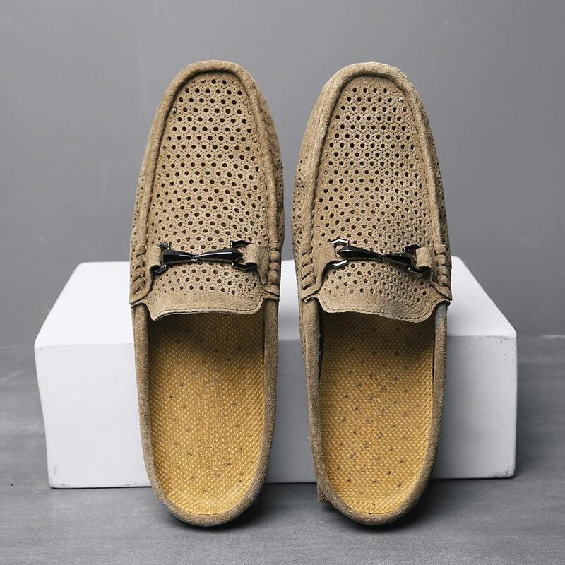 A pair of light brown breathable loafers with metal bit accents, featuring innovative cushioning, displayed upright on white blocks against a gray background.