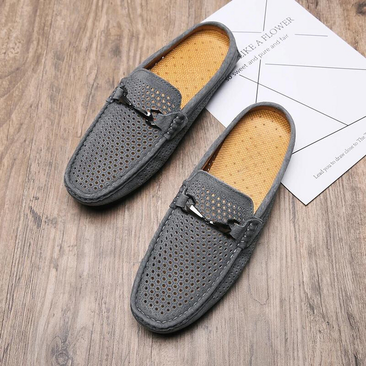 A pair of gray, perforated breathable loafers with tan insoles are placed on a wooden floor. One shoe rests partially on top of a white paper featuring light geometric lines and text.