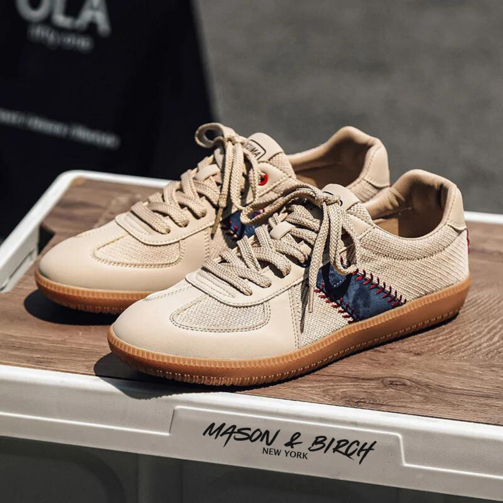 A pair of lightweight, breathable sneakers with gum soles and navy blue-red stitched accents—baseball inspired shoes—are displayed on a wooden surface. The label below reads Mason & Birch New York.