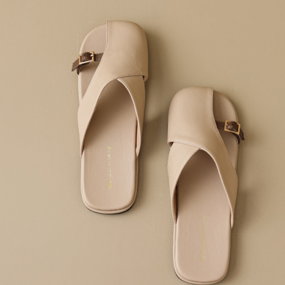 A pair of beige chic slides with square toes, crisscross straps, and small brown buckles, perfect for elevating your summer wardrobe, placed on a matching beige background.