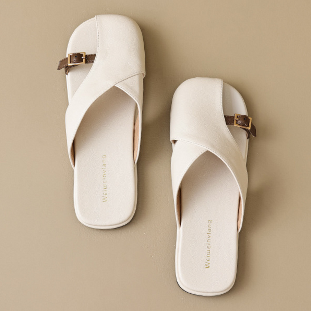 A pair of off-white, slip-on leather slides with crisscross straps and brown buckles, displayed on a beige background—an effortless addition to your summer wardrobe.