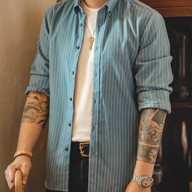 A person wearing a blue pinstripe shirt over a white t-shirt, accessorized with a watch, belt, bracelet, necklace, and displaying tattooed forearms, stands holding a wooden chair. The head is not visible.