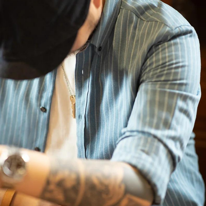 A person in a blue pinstripe shirt made from fine weave cotton, white t-shirt, and black cap looks downward. Their tattooed forearm, wristwatch, and rectangular pendant necklace are visible while their face is partially obscured.