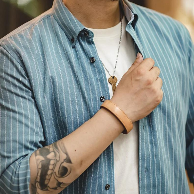 A person wearing a blue pinstripe shirt made of fine weave cotton over a white t-shirt, a key necklace, and a leather bracelet, with a tattoo of a skull on their forearm.