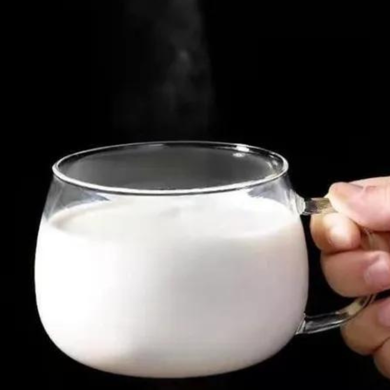 A hand holds a clear glass cup with an ergonomic handle, filled with hot milk against a black background. Steam rises from the heat-resistant glass, emphasizing its warmth and clear design.