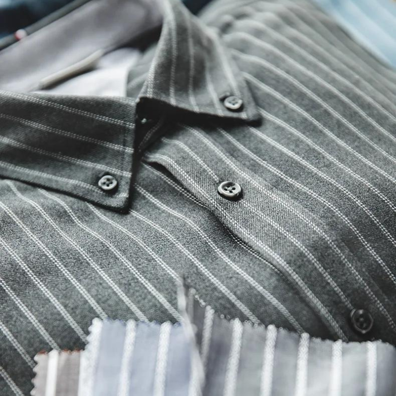 A close-up of a dark gray pinstripe shirt with thin white vertical lines and black buttons, crafted from fine weave cotton. Fabric swatches in various colors and patterns are placed in the foreground.