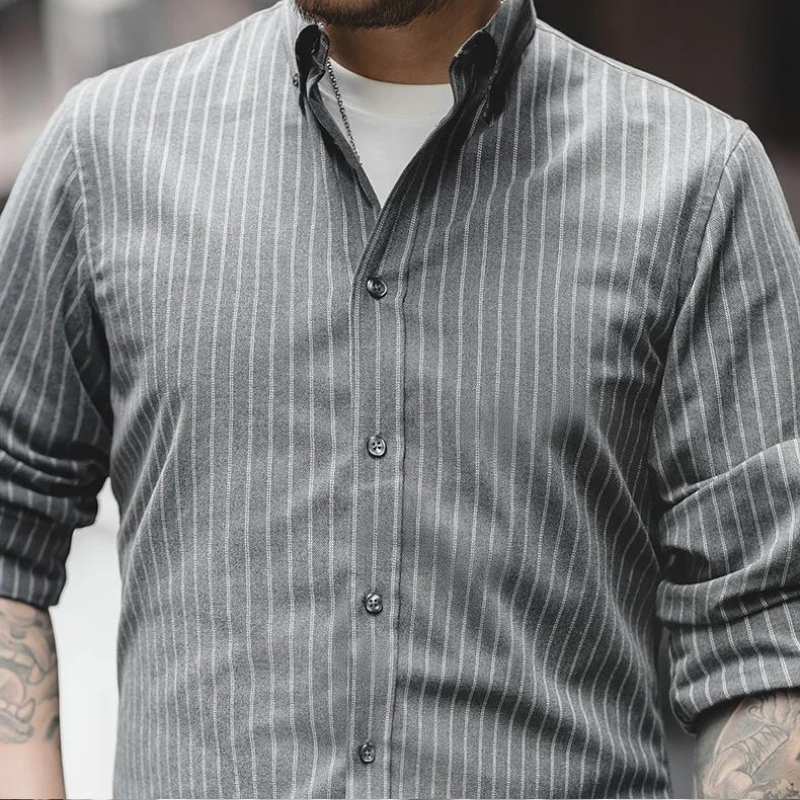 A person wears a gray, wrinkle-resistant pinstripe shirt with white vertical stripes over a white T-shirt. Sleeves are rolled up, revealing tattoos on both forearms. The face is partially out of frame.