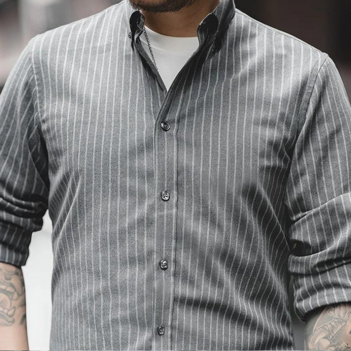 A person wears a gray, wrinkle-resistant pinstripe shirt with white vertical stripes over a white T-shirt. Sleeves are rolled up, revealing tattoos on both forearms. The face is partially out of frame.