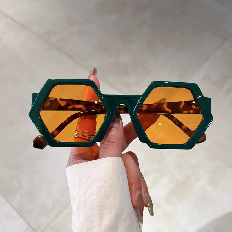 A hand with long, manicured nails holds trend-forward green hexagonal frame sunglasses with amber-tinted lenses and UV400 protection, against a light-colored tiled background.