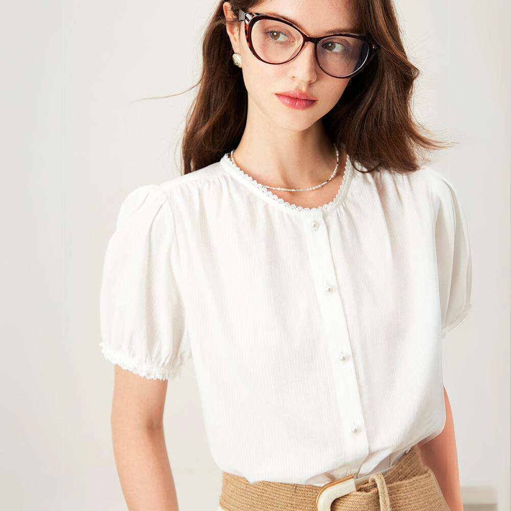 A woman with long brown hair wearing glasses, a white short-sleeved button-up blouse with pearl buttons, a pearl necklace, and tan high-waisted pants stands against a plain light background.