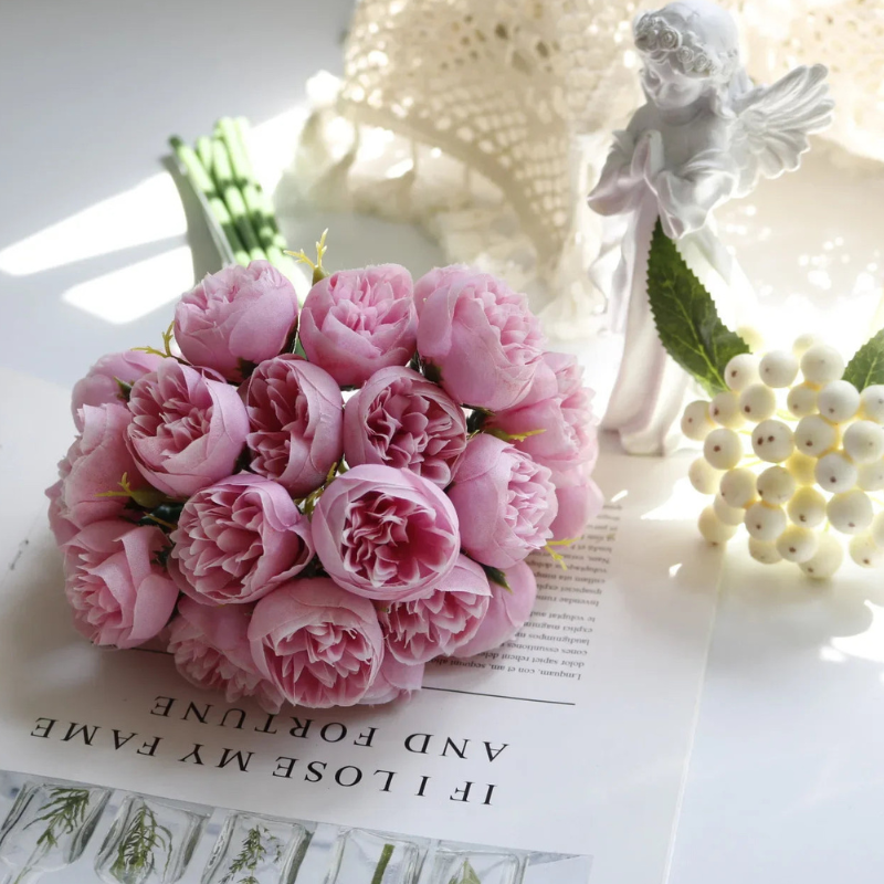 A bouquet of pink peonies with lifelike faux petals rests on an open book. Nearby, a white ceramic angel figurine, white berries, and a lace doily create a charming, maintenance-free display on a bright white surface.