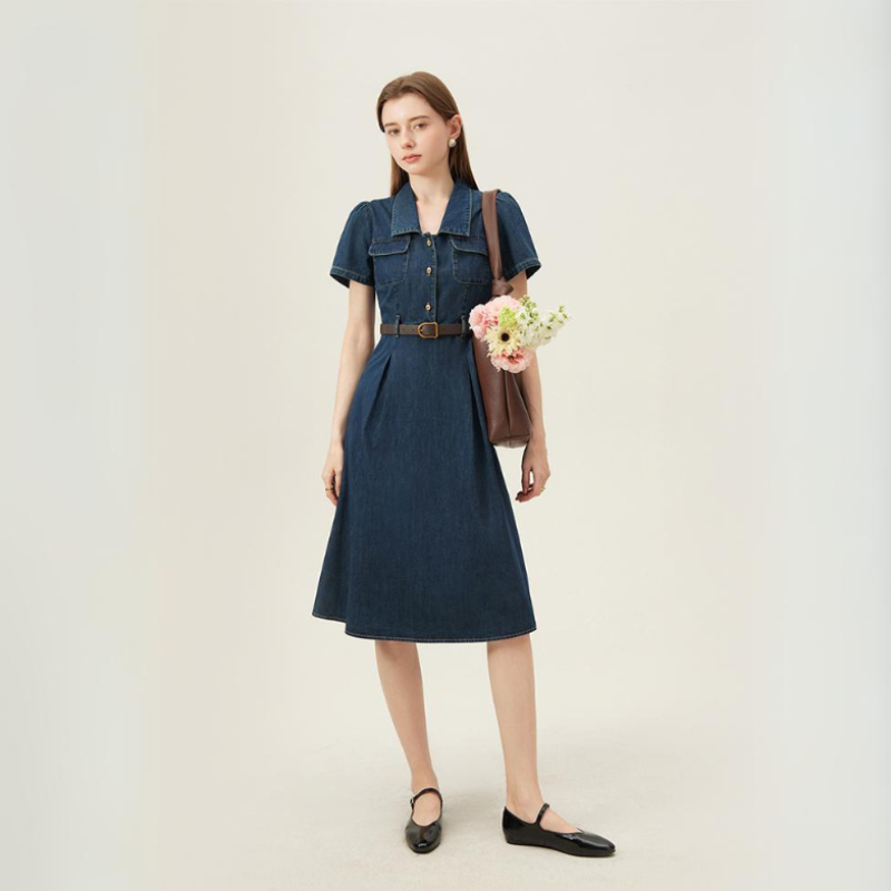 A woman stands against a plain background wearing a belted, short-sleeve denim dress—perfect for summer wear—with black flats. She holds a brown tote bag containing a bouquet of flowers and looks ahead with a neutral expression.